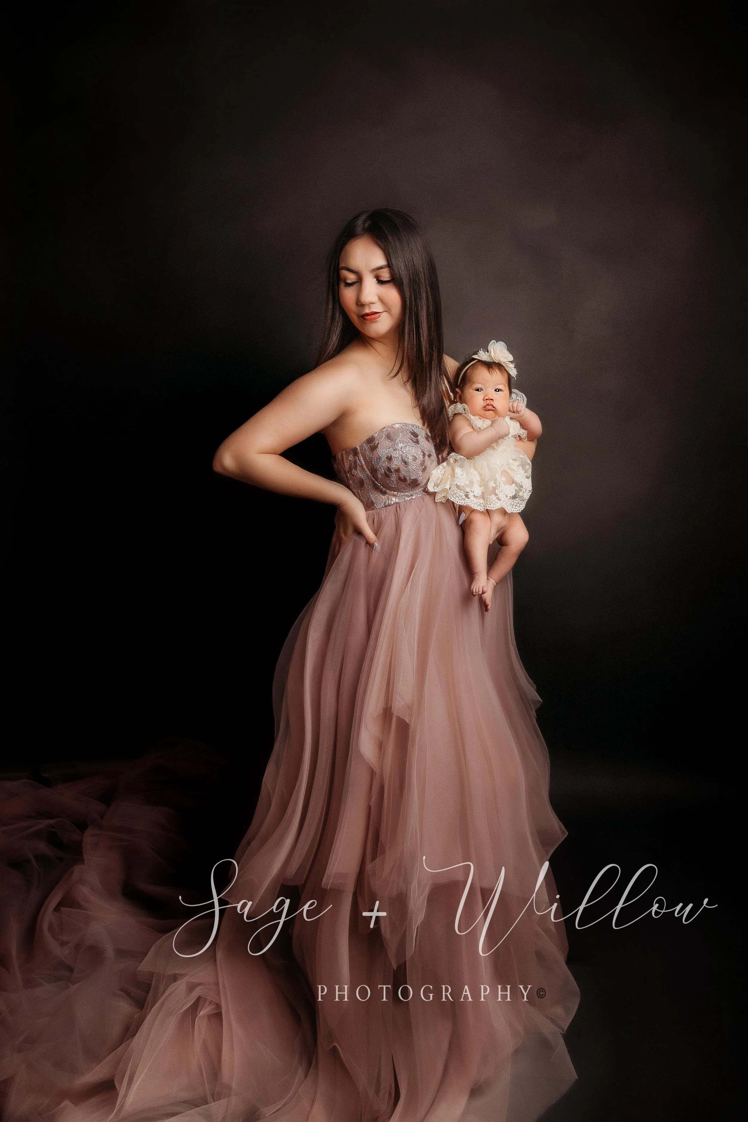 A woman with long dark hair wearing a pink strapless dress with lace details on the bodice, holding a baby girl dressed in a cream-colored lace dress and a matching headband with a flower, against a dark background. The woman is looking down with a gentle expression, and the baby is looking at the camera. The text in the image reads "Sage + Willow Photography."
