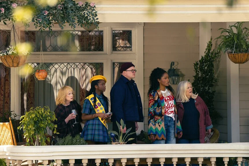 The cast of Peacock’s The ’Burbs, led by Keke Palmer, stands on a suburban porch as neighbors react in shock, capturing the mystery comedy’s suspenseful tone and ensemble cast energy.