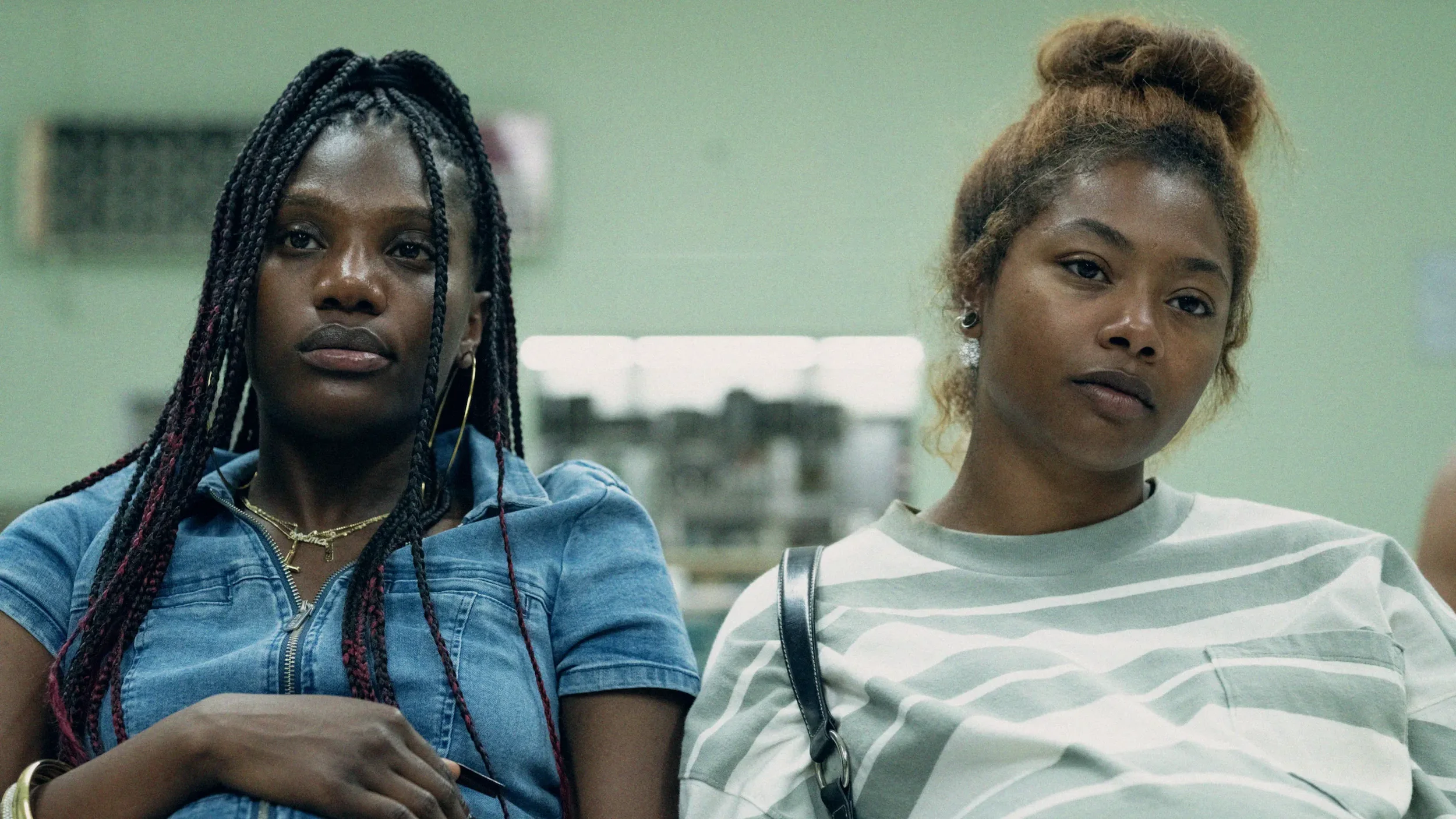 Two young women sit side by side in a waiting room or meeting space, facing forward with thoughtful, serious expressions.
