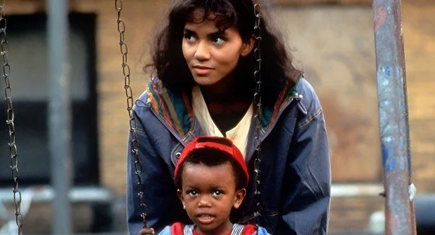 A woman stands behind a small child seated on a playground swing, both looking off-camera as they share a quiet moment outdoors.