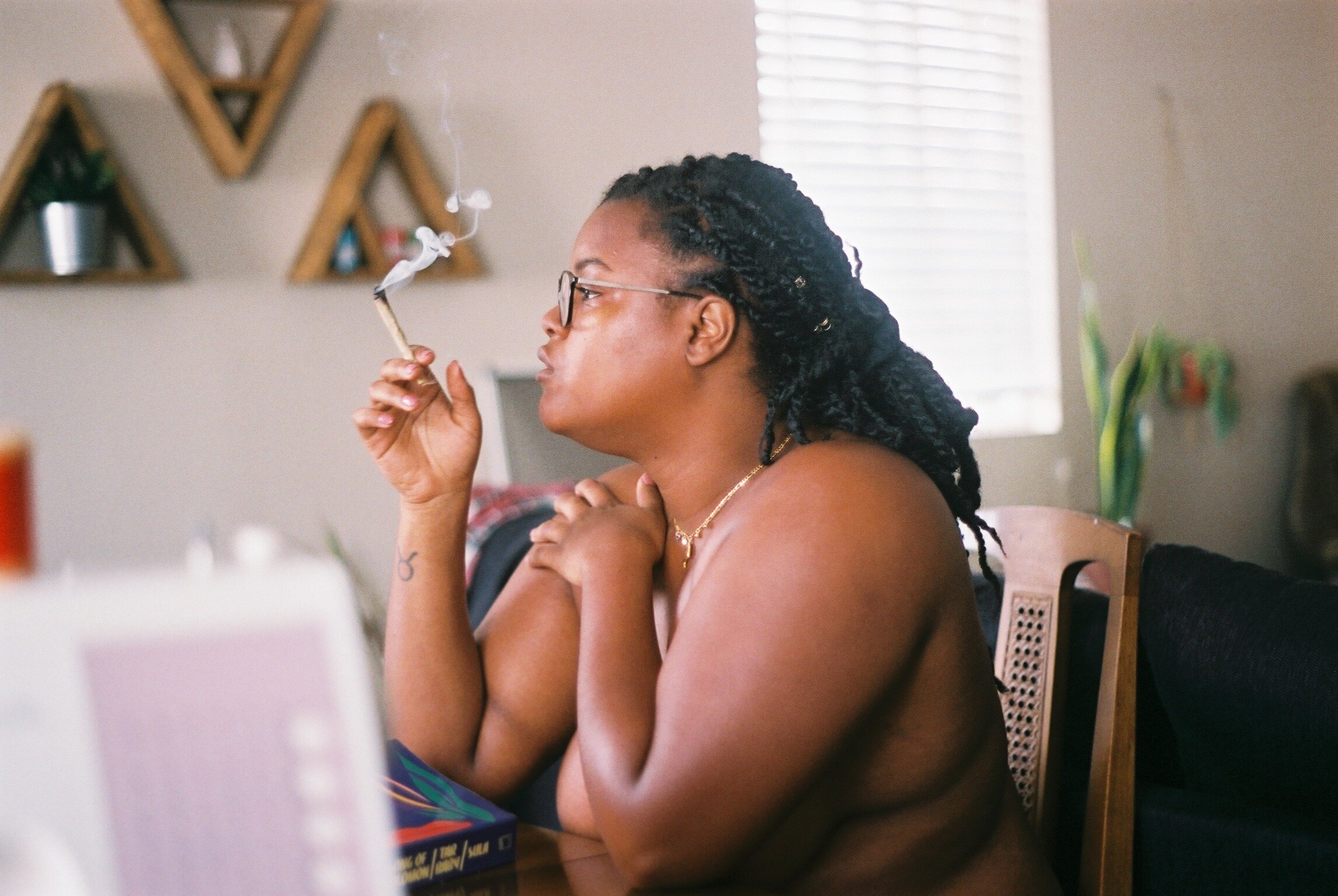 A person with braided hair and glasses sits at a table indoors, holding and smoking a lit joint, with smoke rising as they look off to the side.