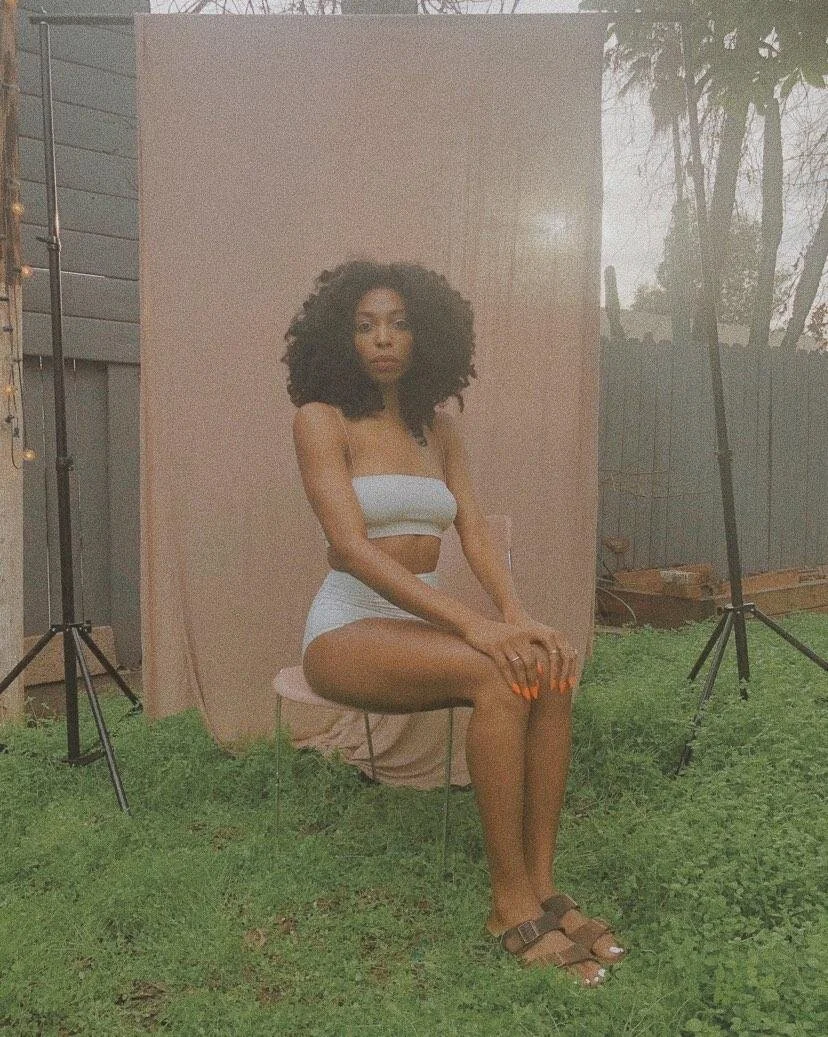 A Black woman is sitting in front of a sheet outside in the grass. She has black curly hair and is wearing a white underwear set.