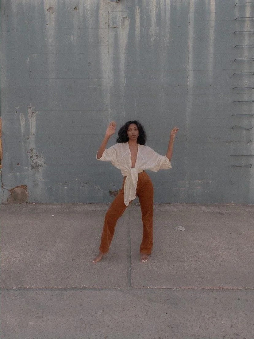 A Black woman is barefoot wearing brown pants and a sheet white shirt. She has long black hair standing in front of a blue wall.