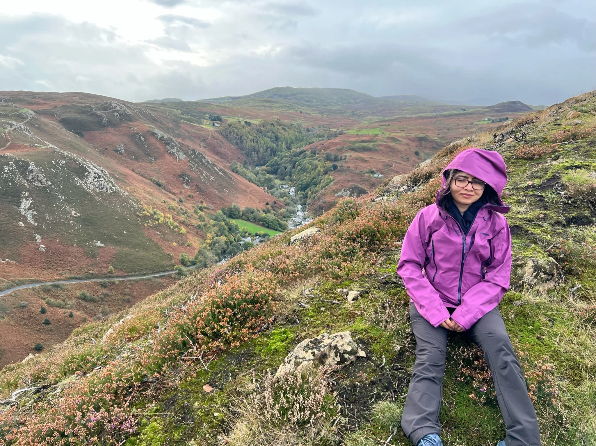 Malala Yousafzai in a purple jacket sits on a grassy hillside overlooking a sweeping valley of rolling hills, winding roads, and autumn-toned landscapes beneath a cloudy sky.