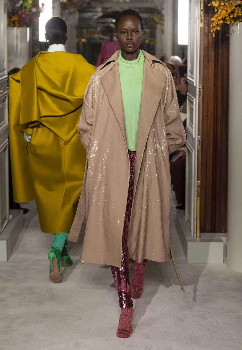 A model walks the runway in a modern Valentino look featuring a sheer beige trench layered over a mint knit and shimmering sequin trousers, highlighting the house’s mastery of unexpected color pairings and refined contemporary elegance.