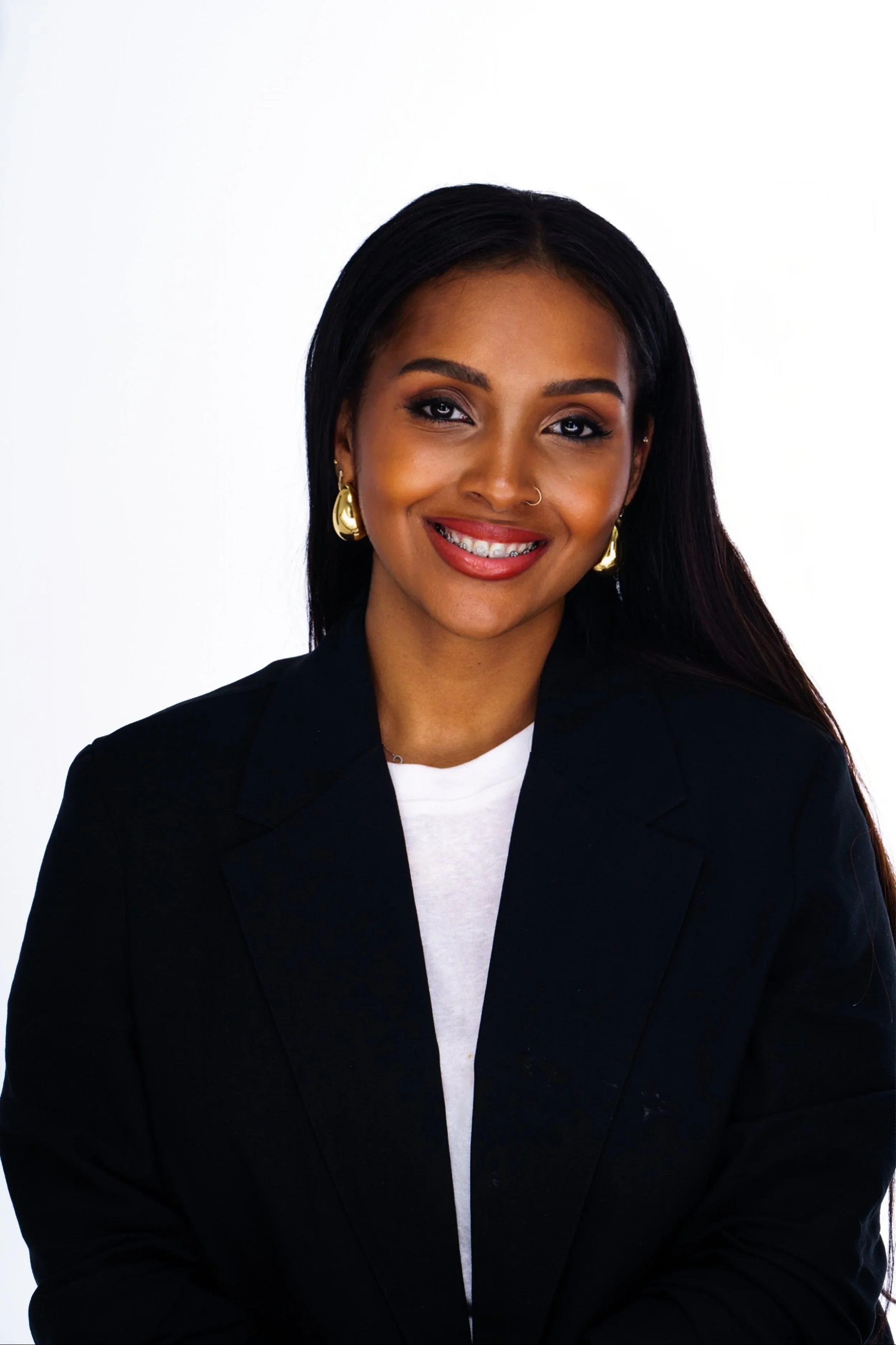 A Black woman with long black hair is wearing a black blazer over a white t-shirt and smiling on a white background