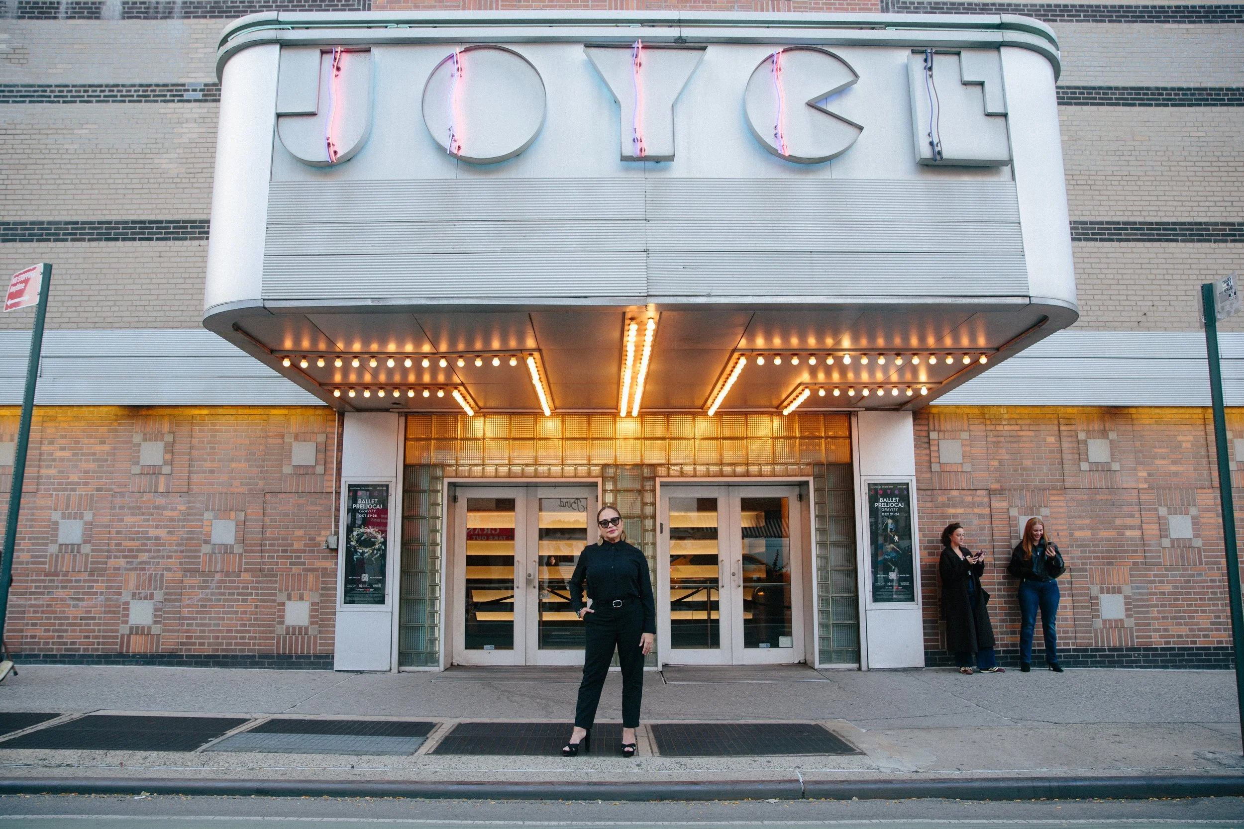 A woman dressed in black stands confidently in front of a vintage theater entrance with a glowing marquee that reads “JOYCE,” while two people linger near the doorway.