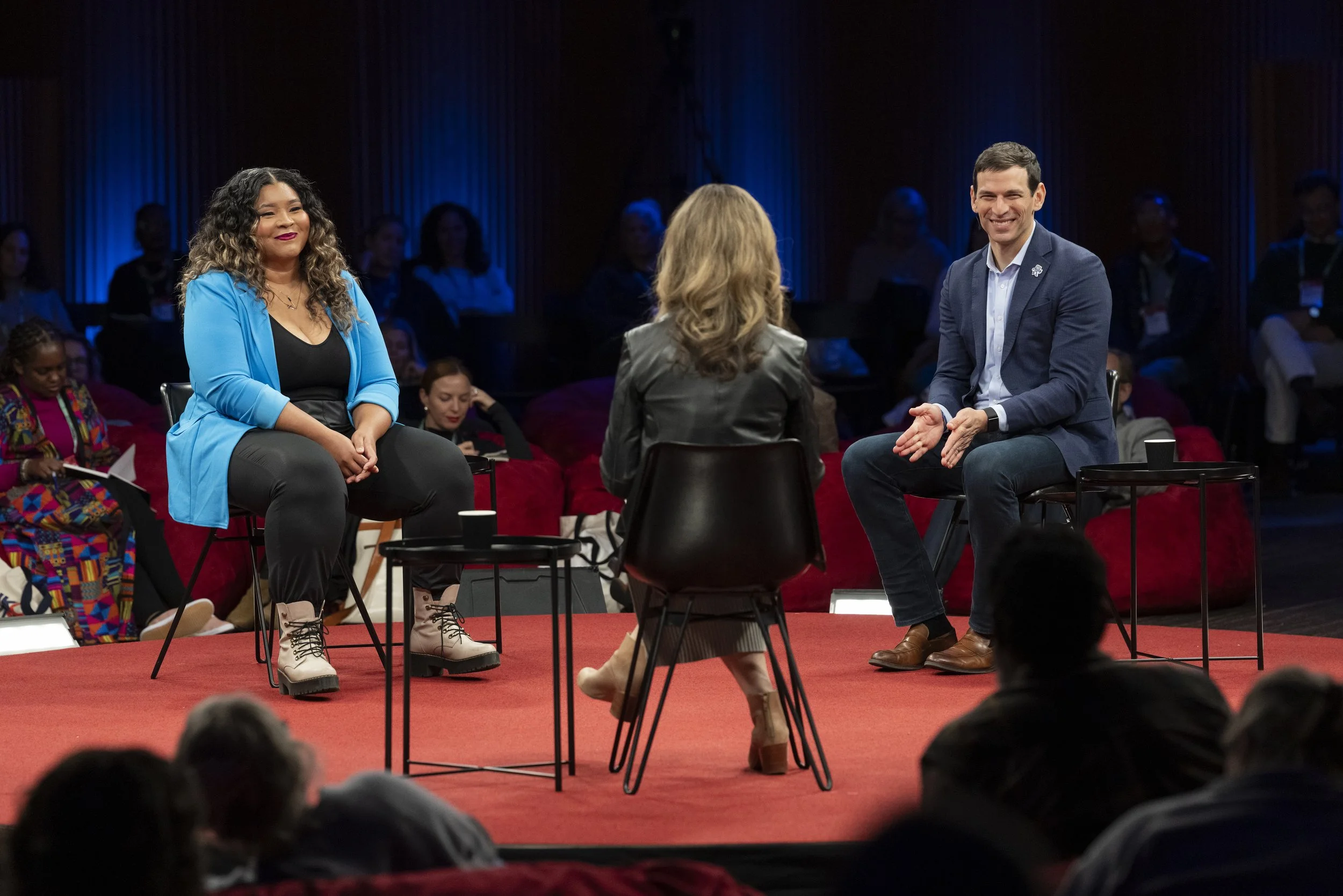 Three speakers participate in a lively panel discussion on a red stage, engaging an attentive audience in a dynamic, conference-style conversation.
