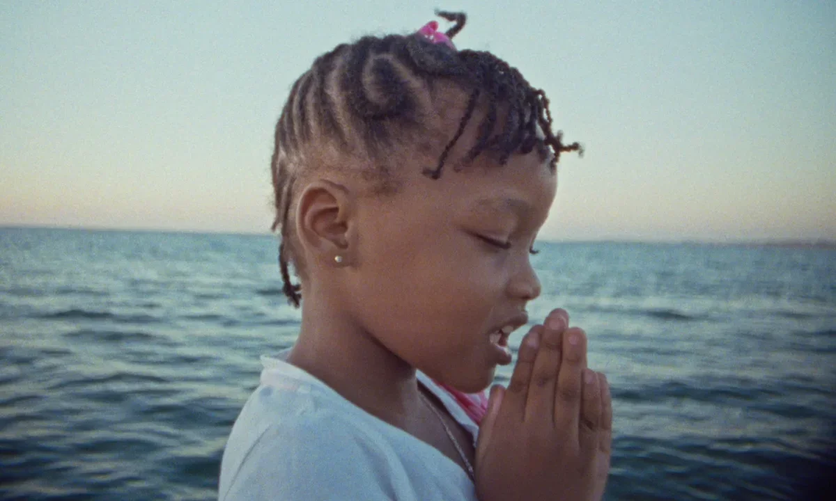 A young girl stands beside a large body of water with her eyes closed and hands clasped together, framed by a soft sky and calm horizon.