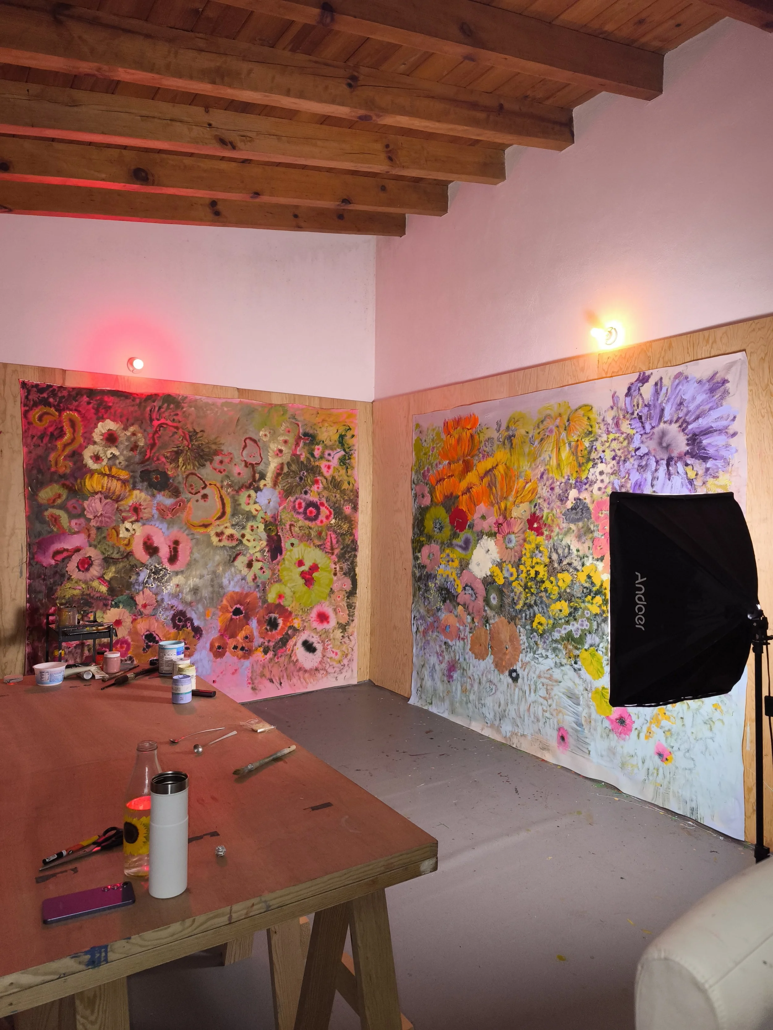 Inside a warm, wood-beamed studio, two monumental floral canvases glow against the walls beside a long worktable scattered with brushes, jars, and tools, capturing the immersive environment of her practice.