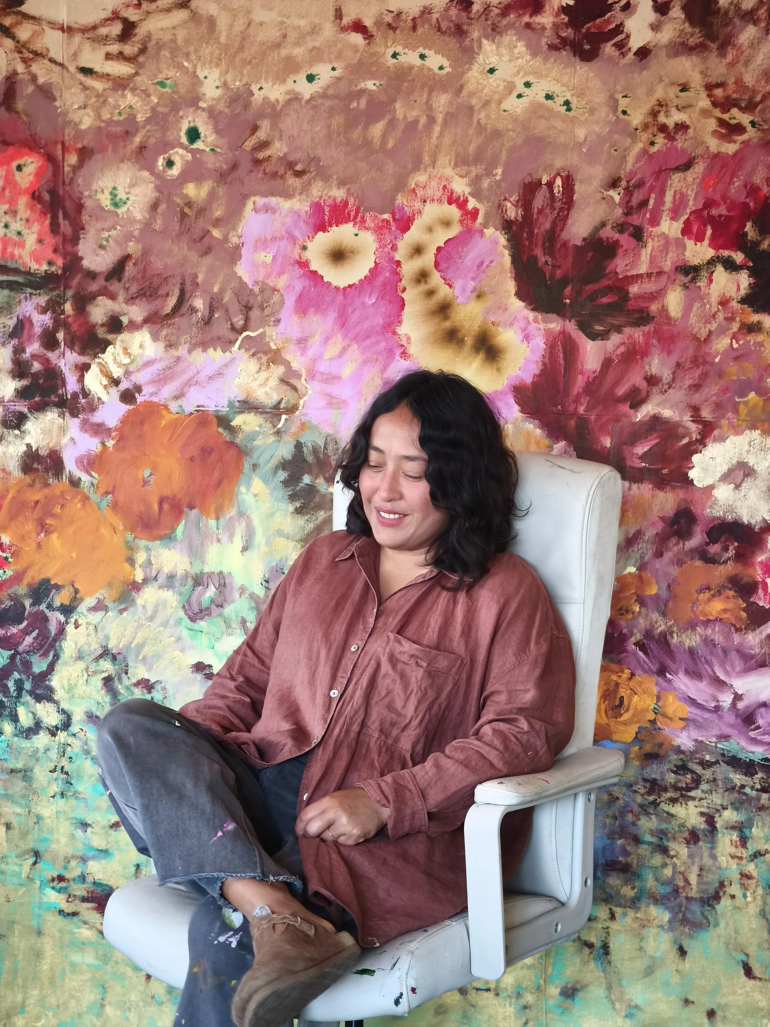 The artist sits relaxed in a white studio chair before a large, expressive floral painting in pink, ochre, and mossy green, her paint-splattered clothes echoing the lush, tactile surface behind her.