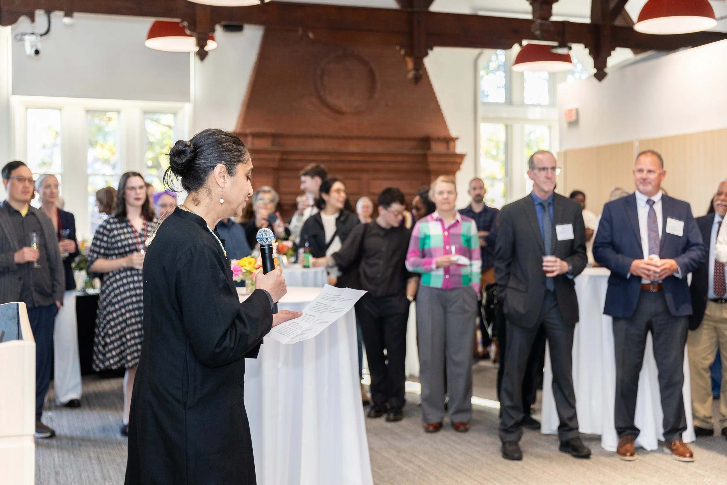 A speaker addresses a large audience at a formal reception, delivering remarks to a diverse crowd gathered in a bright event space for a celebratory gathering.