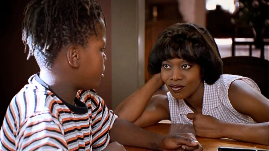 A woman rests her head on her hand as she gazes attentively at a young child seated across from her at a table during an intimate conversation.