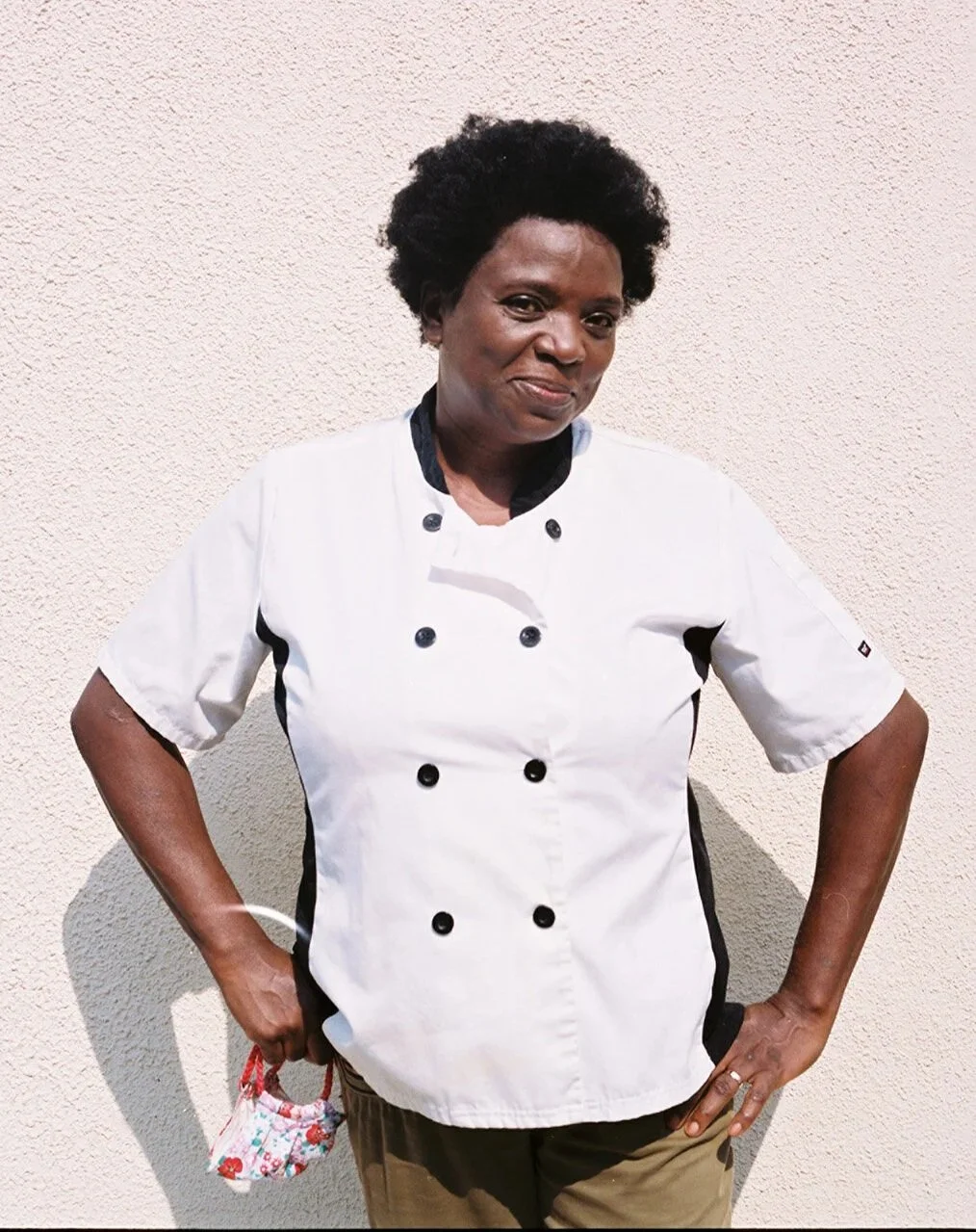 A Black woman with short black hair smiles off into the sun. She stands in front of a white wall wearing a chefs coat.
