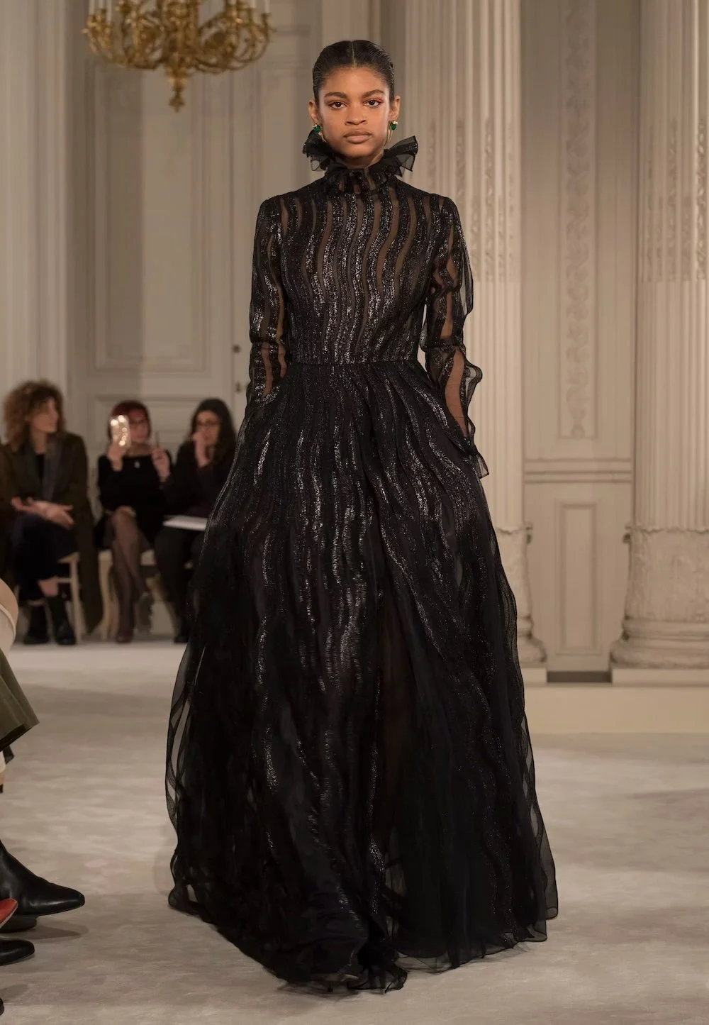 A model commands the runway in a flowing black Valentino gown with sheer, subtly shimmering layers and a high neckline, embodying the house’s timeless elegance and quietly dramatic couture sensibility.