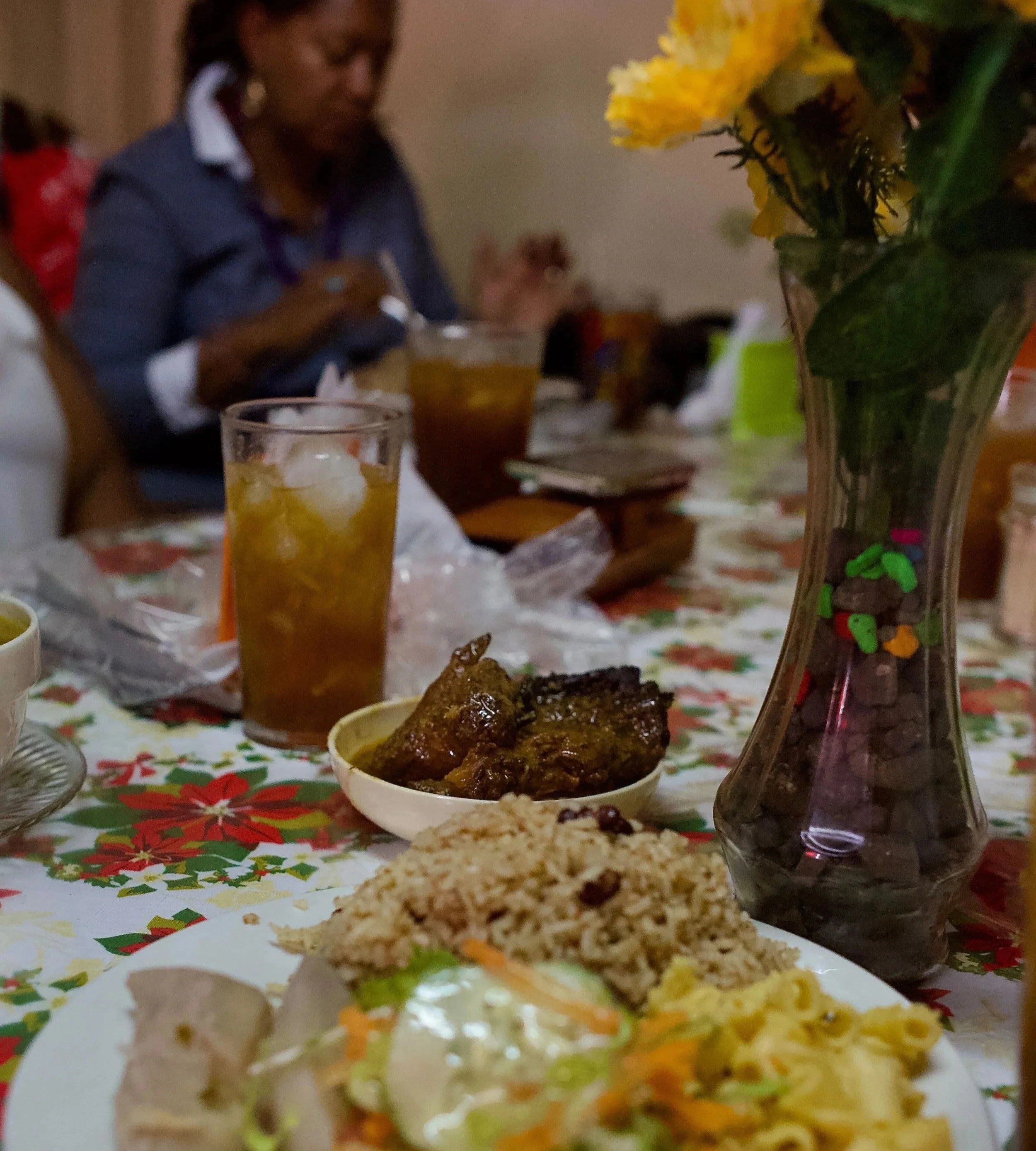 A plate of food and glass of tea sitting on a table next to flowers and a woman is eating in the background.