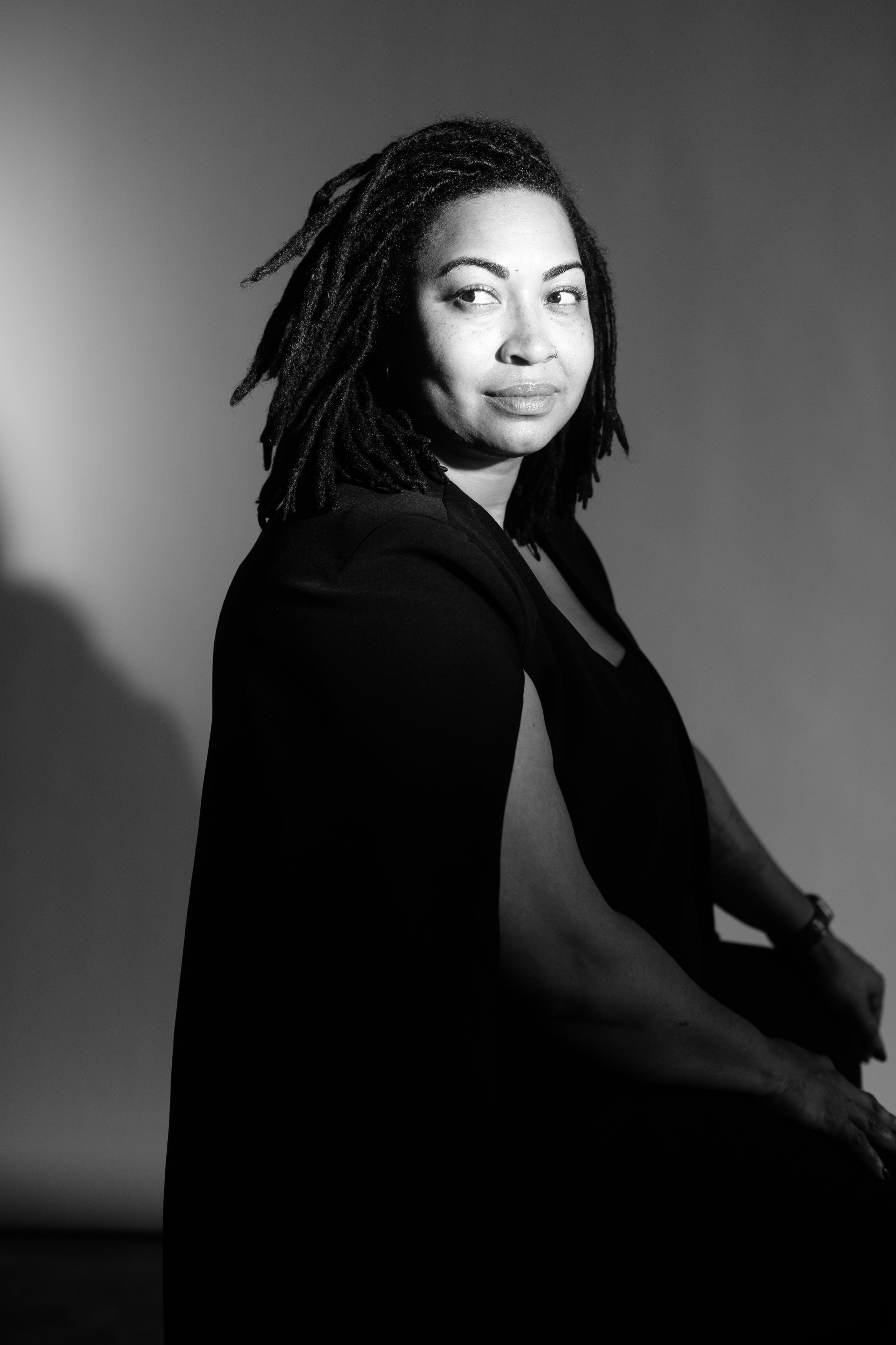 A woman wearing a black cape and dreaded hair looks off to the side with a slight smile against a white background.