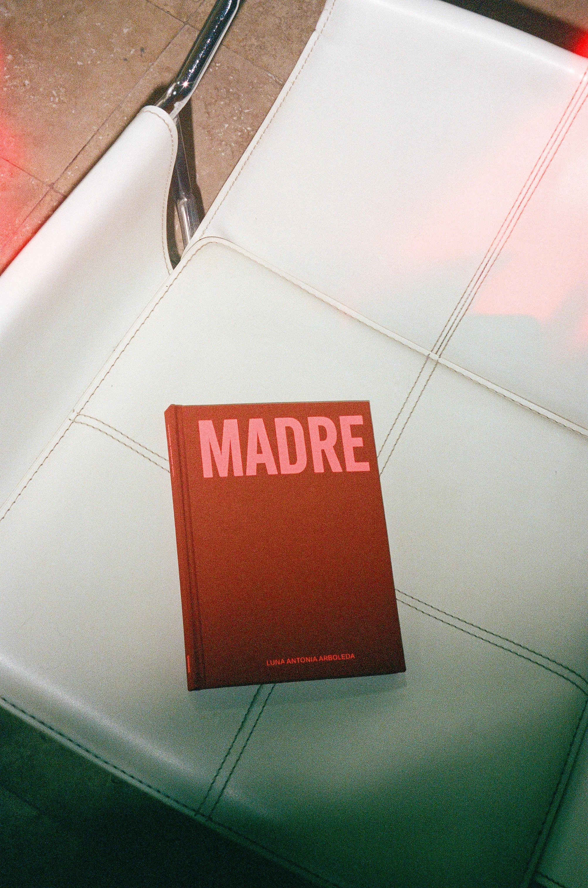 Cover of Madre — A deep red hardcover copy of Madre by Luna Antonia Arboleda rests on a white leather chair, photographed with soft analog light leaks and warm tones.