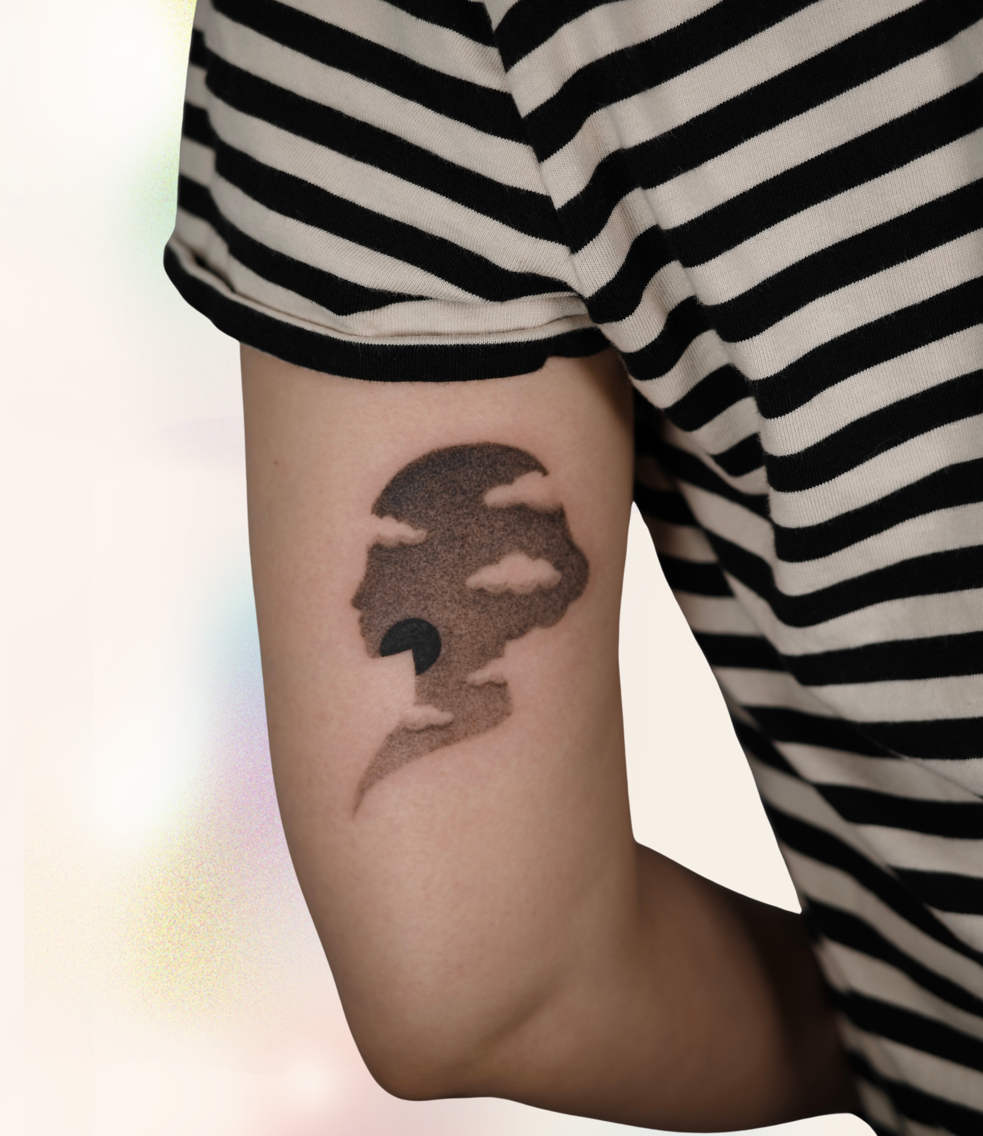 A person displays a stylized black-and-gray tattoo on their upper arm featuring a silhouetted face formed by clouds and soft dotwork shading.