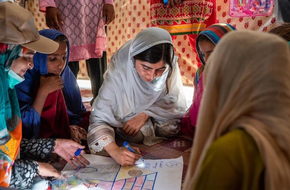 Malala Yousafzai and young girls sit together indoors, collaborating on a colorful drawing as one person sketches while others watch closely.