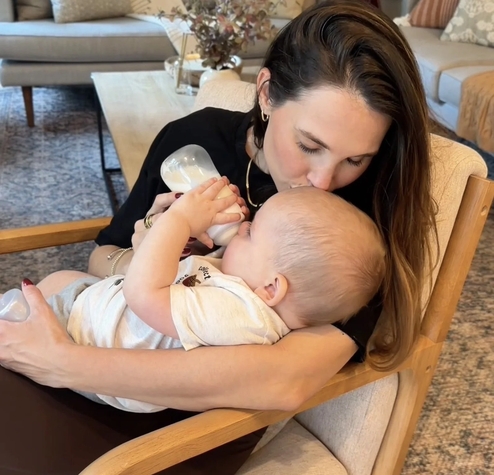 A woman is feeding her baby a bottle and kissing the baby's head in a living room.
