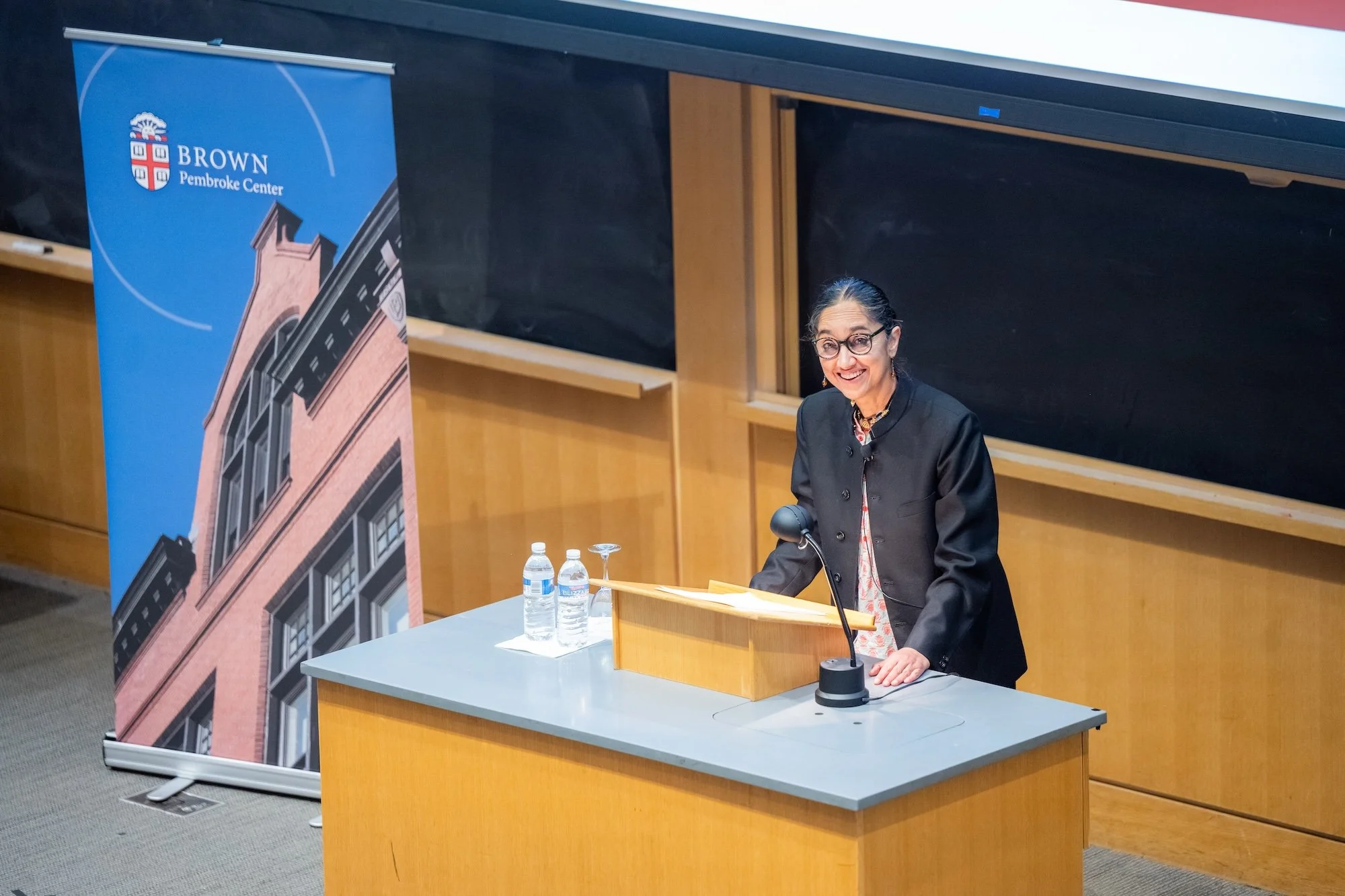 A distinguished speaker delivers a lecture at Brown University’s Pembroke Center, standing at a podium in a large academic auditorium with a welcoming, scholarly atmosphere.