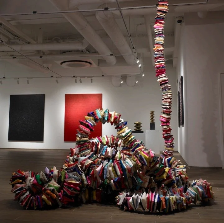 A large-scale gallery installation composed of hundreds of brightly colored pillows cascades across the floor and rises in a towering vertical column, set against minimalist white walls with contemporary paintings in the background.