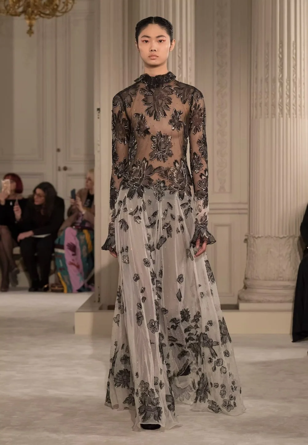 A model glides down the runway in an ethereal Valentino gown featuring sheer layers and intricate black floral embroidery, exemplifying the house’s signature balance of delicate romance and refined couture elegance.