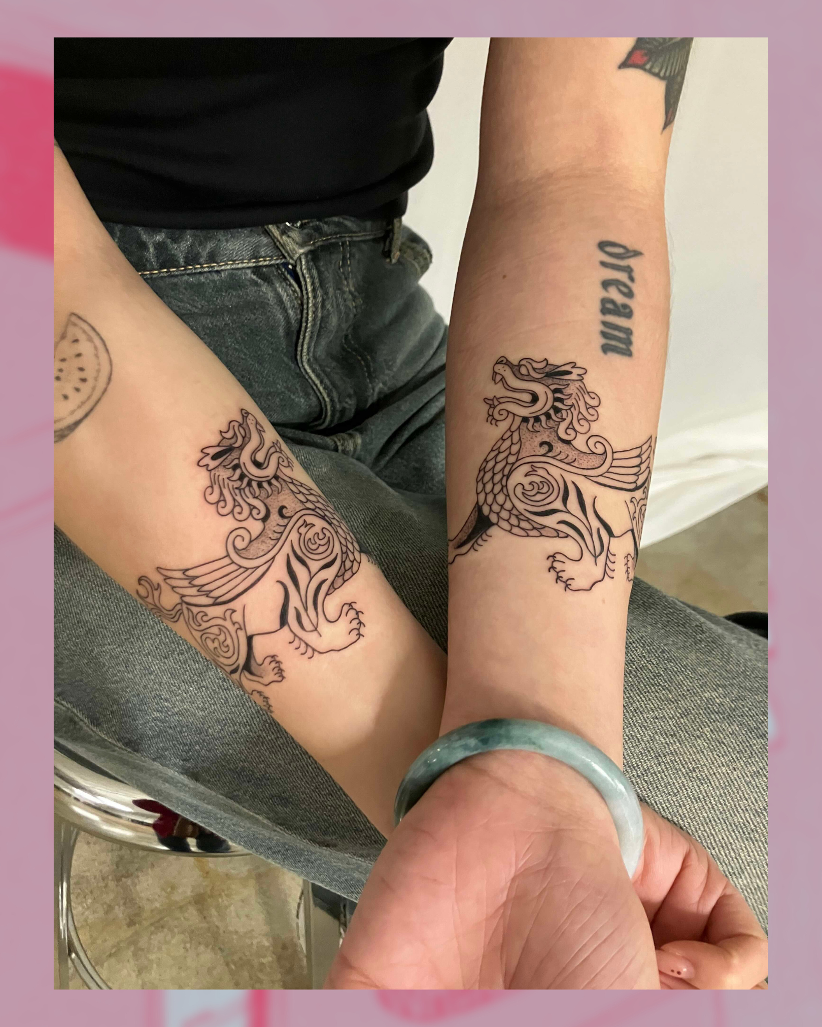 Two forearms display matching black ink tattoos of a stylized winged creature, with the word “dream” tattooed on one arm.