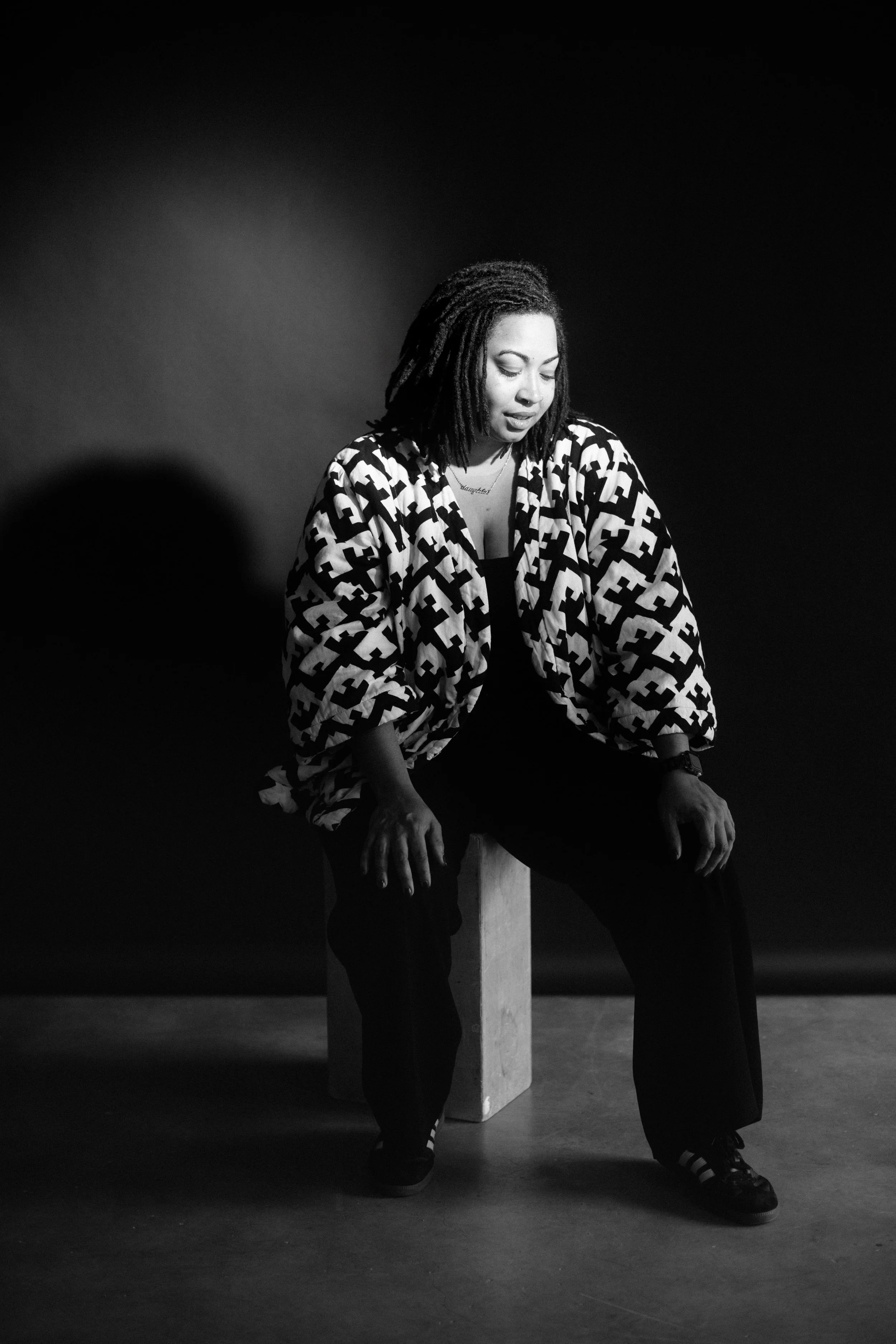 A woman is smiling, sitting on a box wearing a houndstooth jacket in front of a black background.