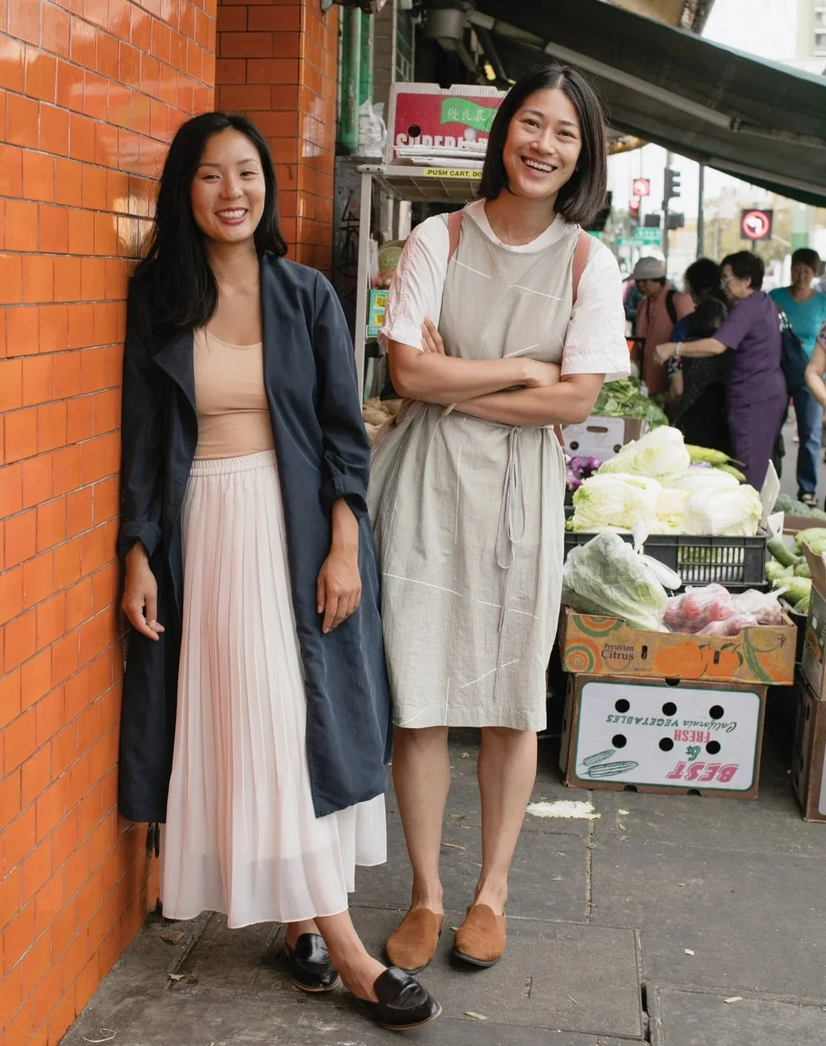 Two Asian women standing on a street next to a brick wall smiling