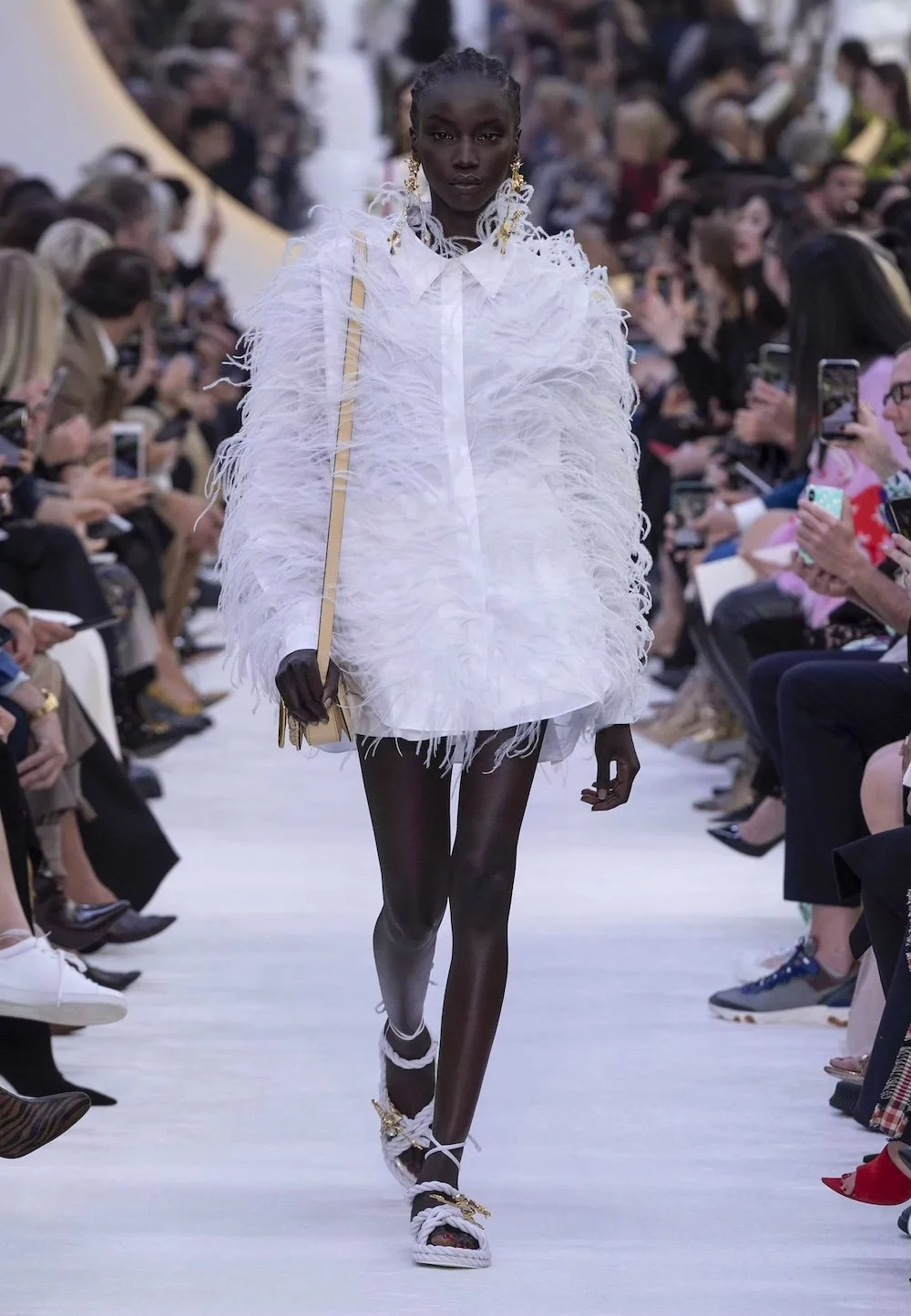 A model walks the runway in a Valentino look featuring a feather-textured white mini dress paired with delicate accessories, reflecting the house’s signature blend of ethereal elegance, modern femininity, and couture craftsmanship.
