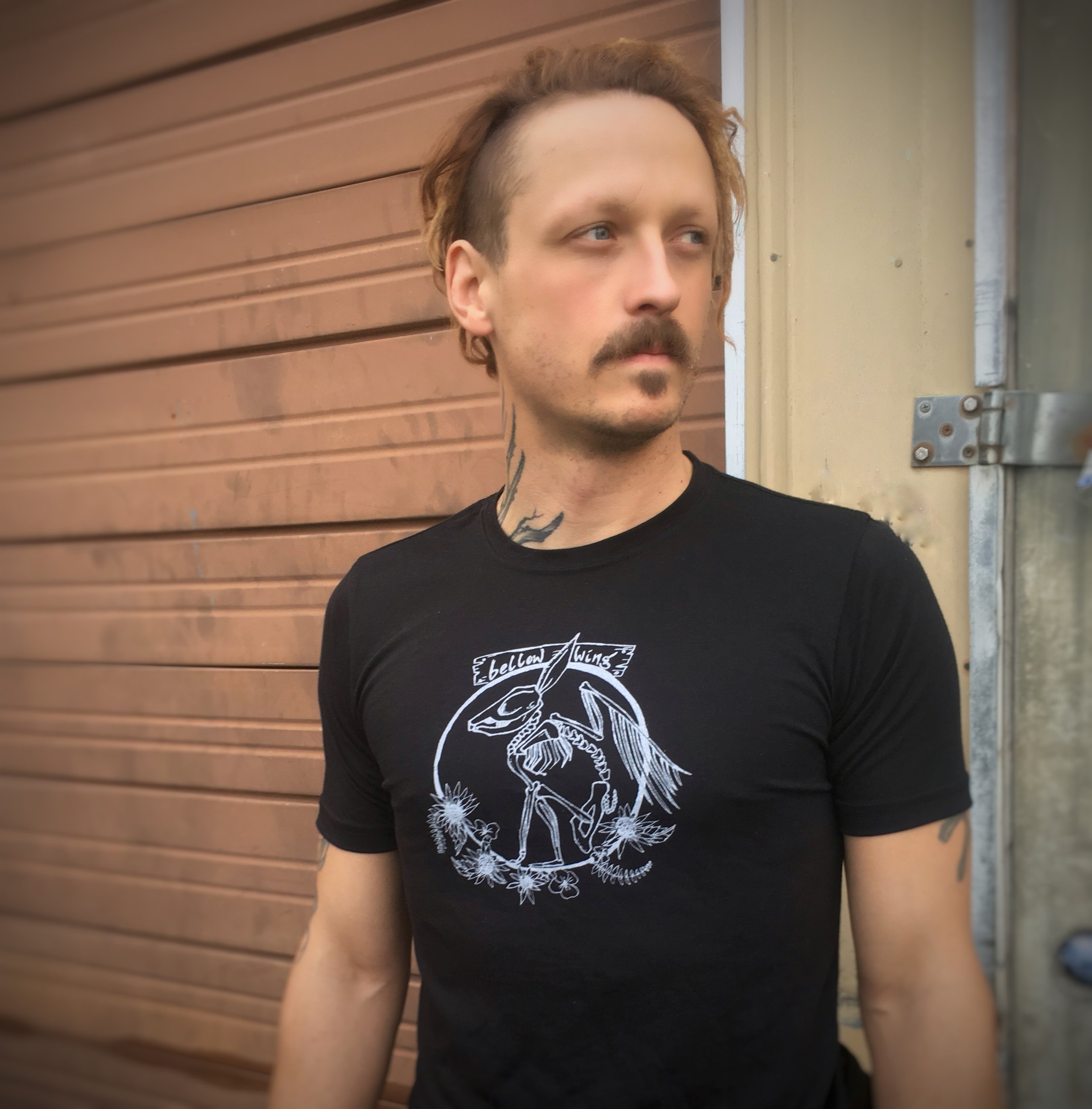 Man wears black unisex t-shirt with Bellow Wing winged rabbit skeleton logo