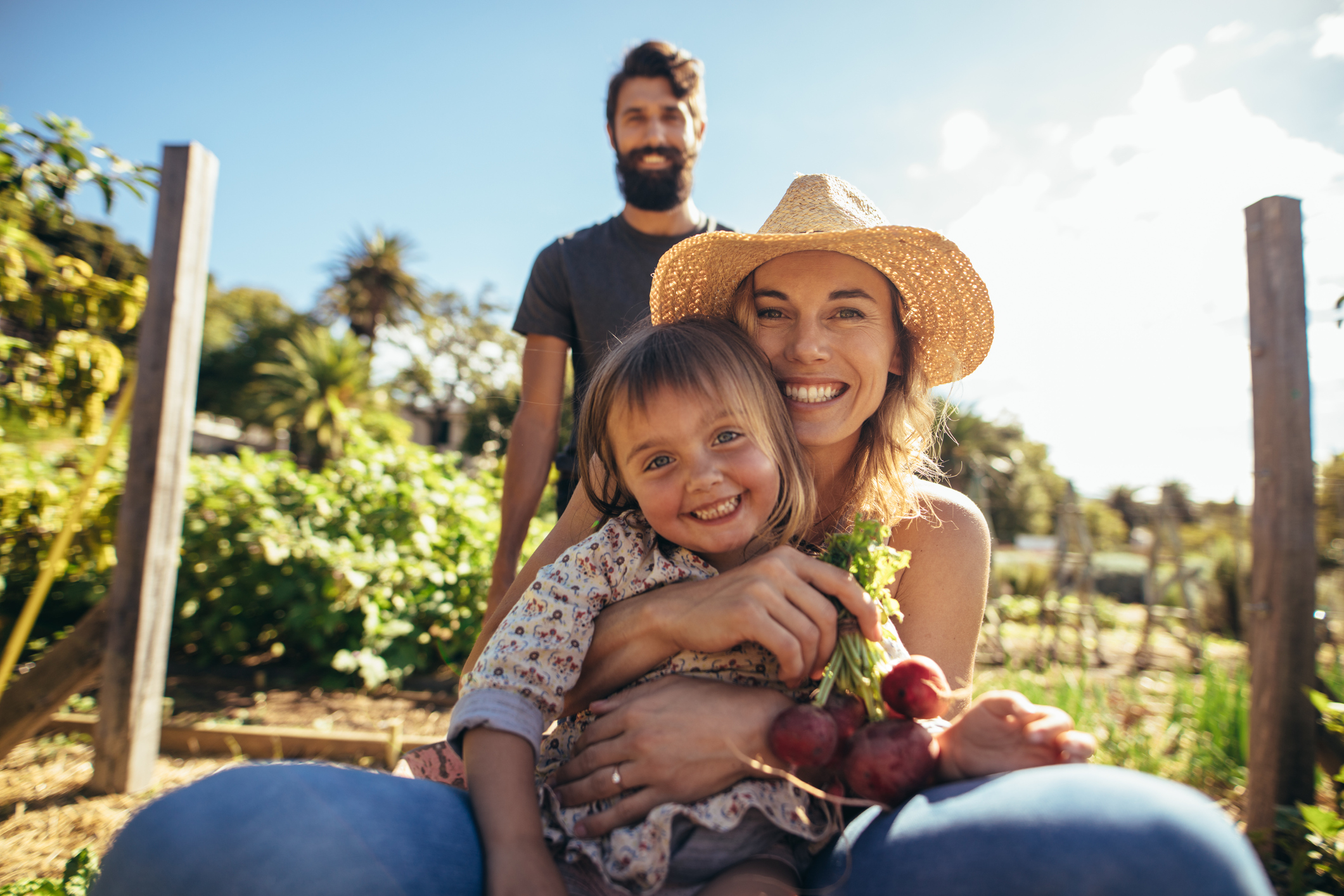 Portrait of beautiful woman with her daughter sitting in wheelbarrow while man pushing. Smiling young mother and daughter enjoying in wheelbarrow ride at outdoors at farm.