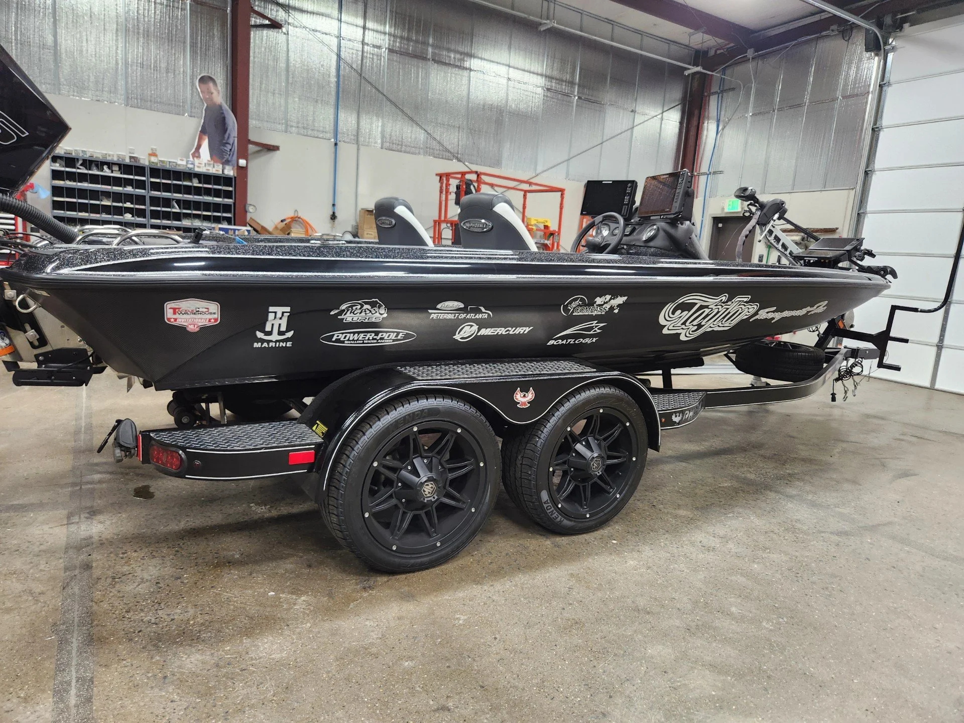 Wrapped Black bass boat on a trailer inside a workshop. The boat has various sponsor logos and a mounted fish finder. The workshop has tools and equipment in the background.