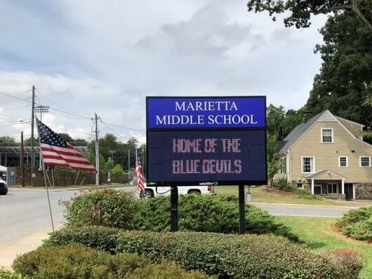Electronic sign at Marietta Middle School displaying 'Home of the Blue Devils' with American flags nearby and residential houses in the background.