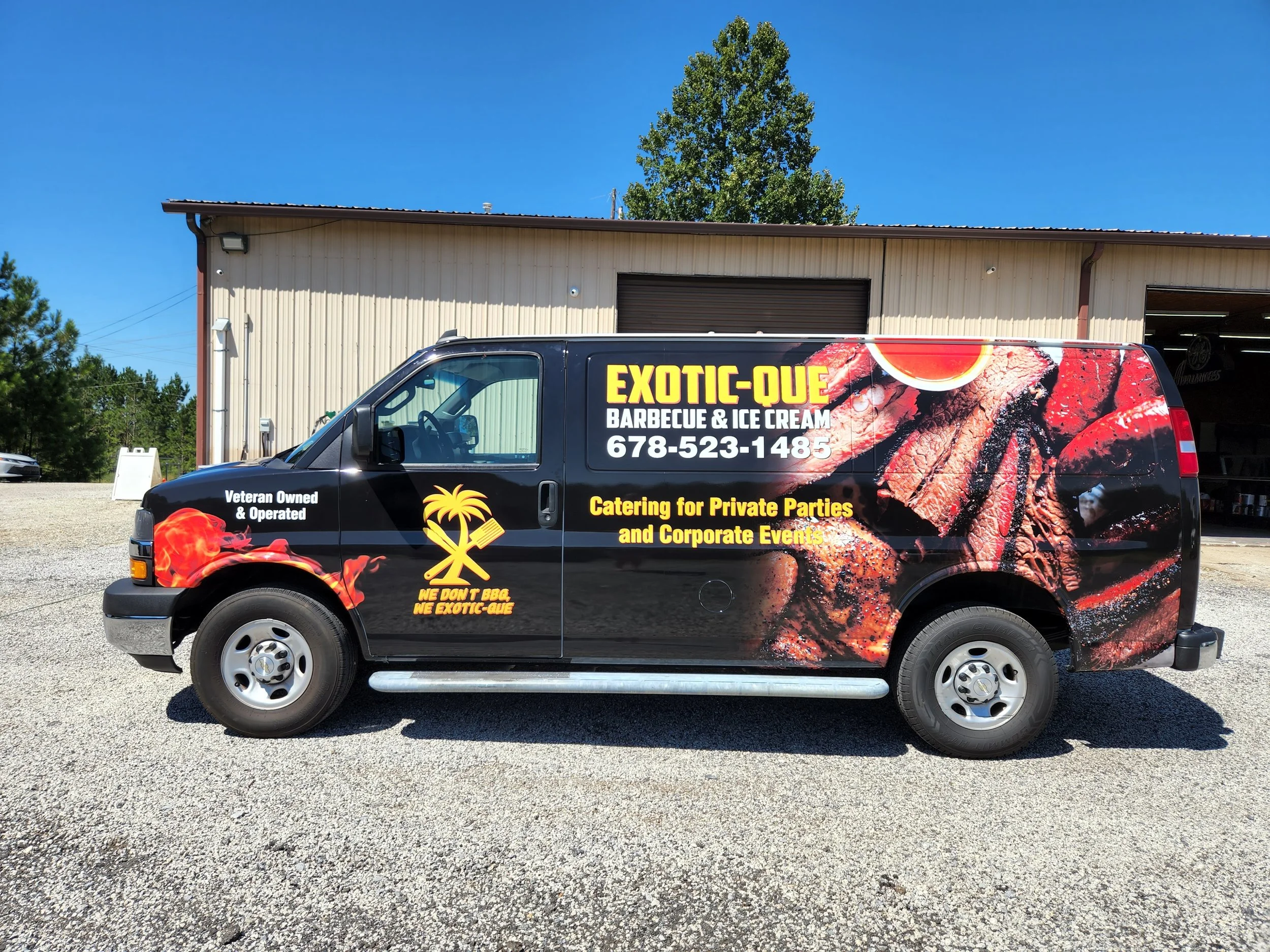 Black cargo van with bright yellow and white lettering advertising Exotic-Que Barbecue & Ice Cream, featuring an image of grilled meat and fire, parked outside a large metal building on a gravel lot with trees in the background.