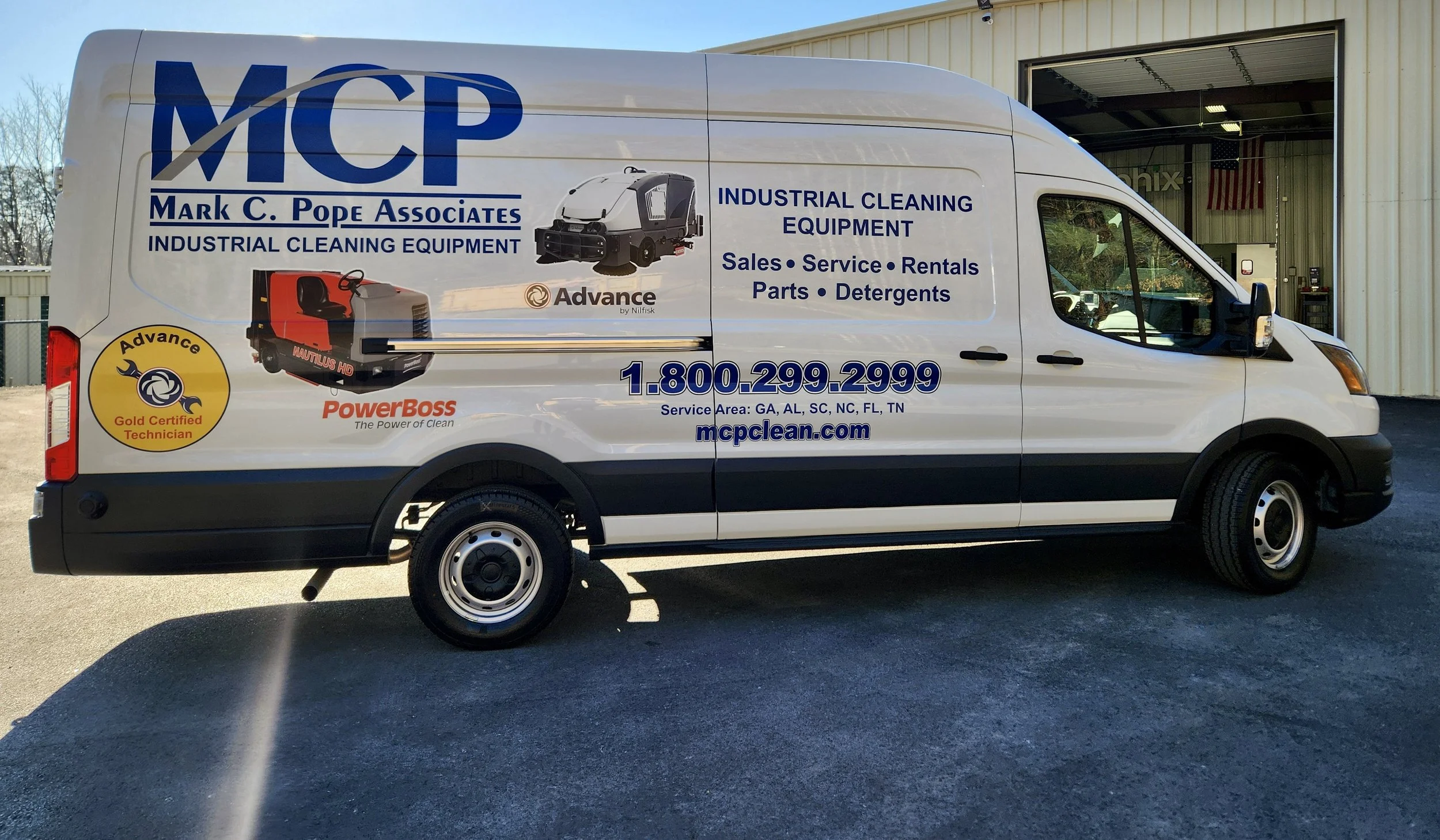 White service van with advertising for McP, Mark C. Pope Associates, with contact number 1-800-299-2999, positioned outside a warehouse with an American flag inside, featuring images of industrial cleaning equipment and logos for Advance and PowerBos