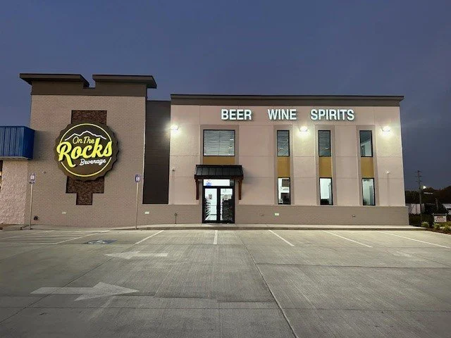 Front of a beverage store called On the Rocks with signage for beer, wine, and spirits, and a large parking lot in front.