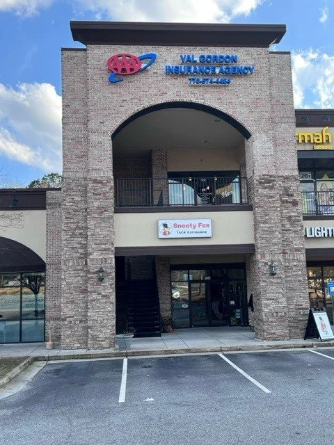 Front view of a two-story commercial building with a sign for Val Gordon Insurance Agency on the upper level and Snooty Fox Tack Exchange on the lower level, with parking spaces in front.