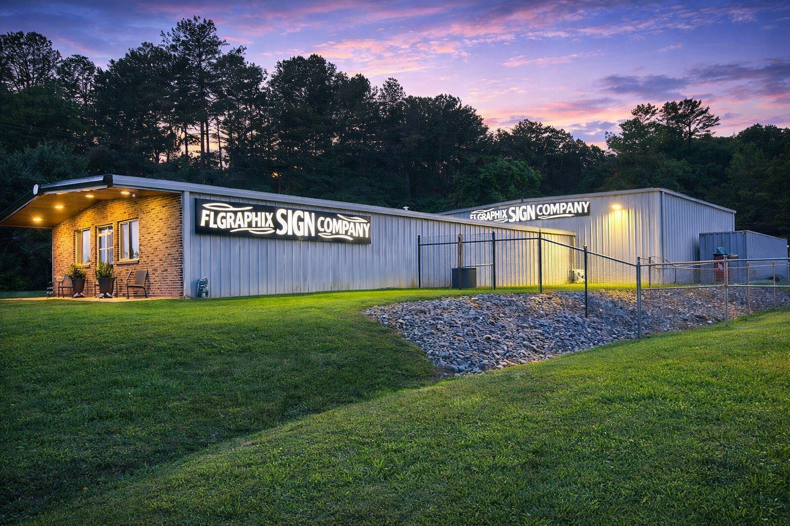 A building with a sign that reads 'FLGRAPHIX SIGN COMPANY' in bright lights at dusk. The building has brick and metal siding, with small windows, outdoor chairs, potted plants, a sloped lawn, and a rocky area in front. Trees are visible in the backgr