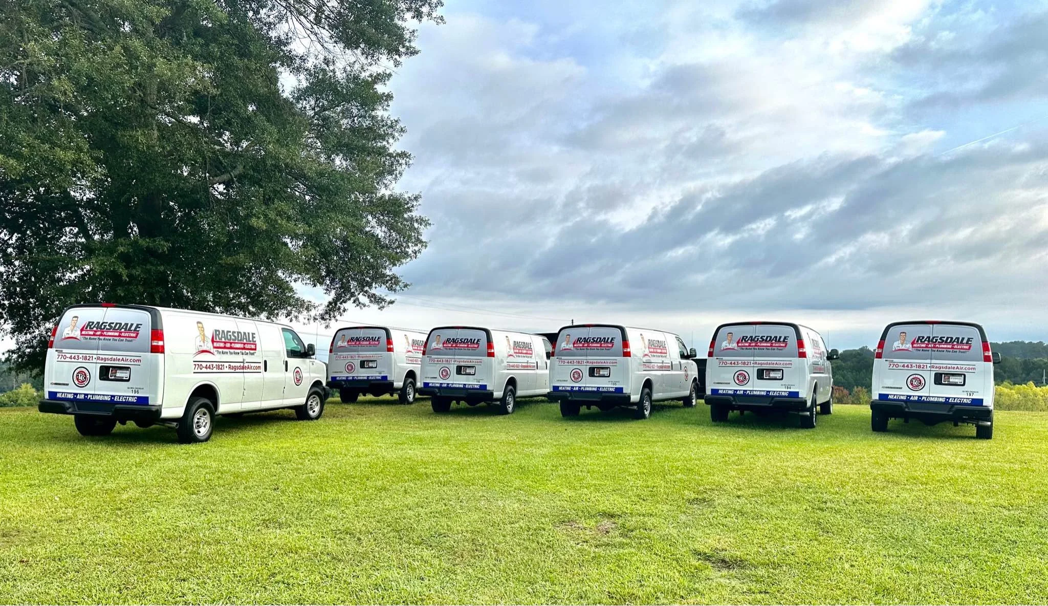 Six white service vans with company branding are parked on a grassy field under a cloudy sky, with a large tree to the left.