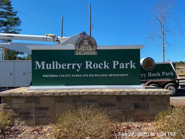 Sign for Mulberry Rock Park with the logo of the Paulding County Parks and Recreation Department, mounted on a stone base with some shrubbery in front.