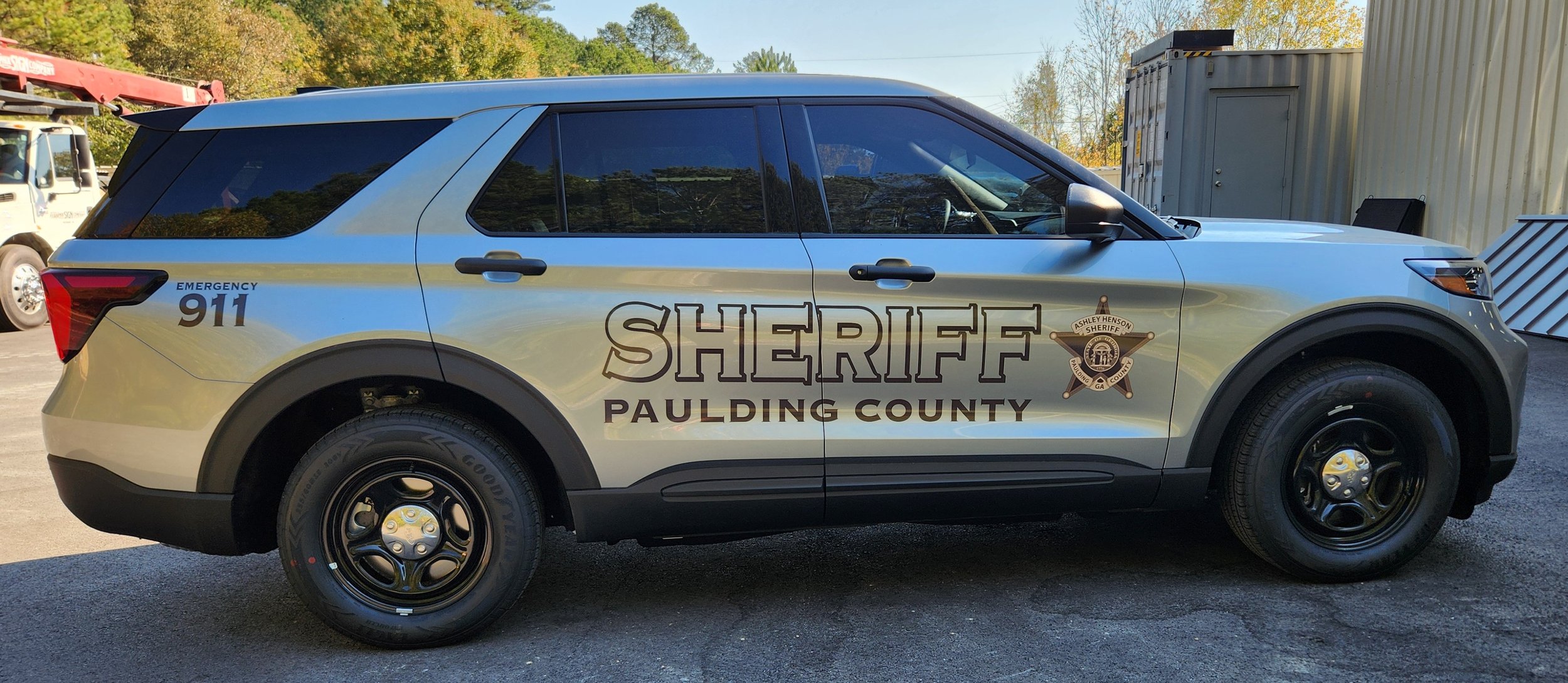 Sheriff's vehicle from Paulding County with Sheriff badge and emergency contact info parked outside near trees and a chain-link fence.
