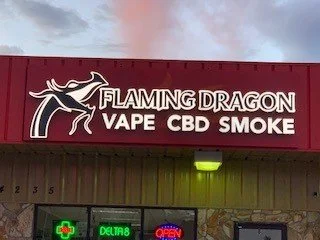 Storefront sign for 'Flaming Dragon Vape CBD Smoke' with a dragon logo and neon signs below.