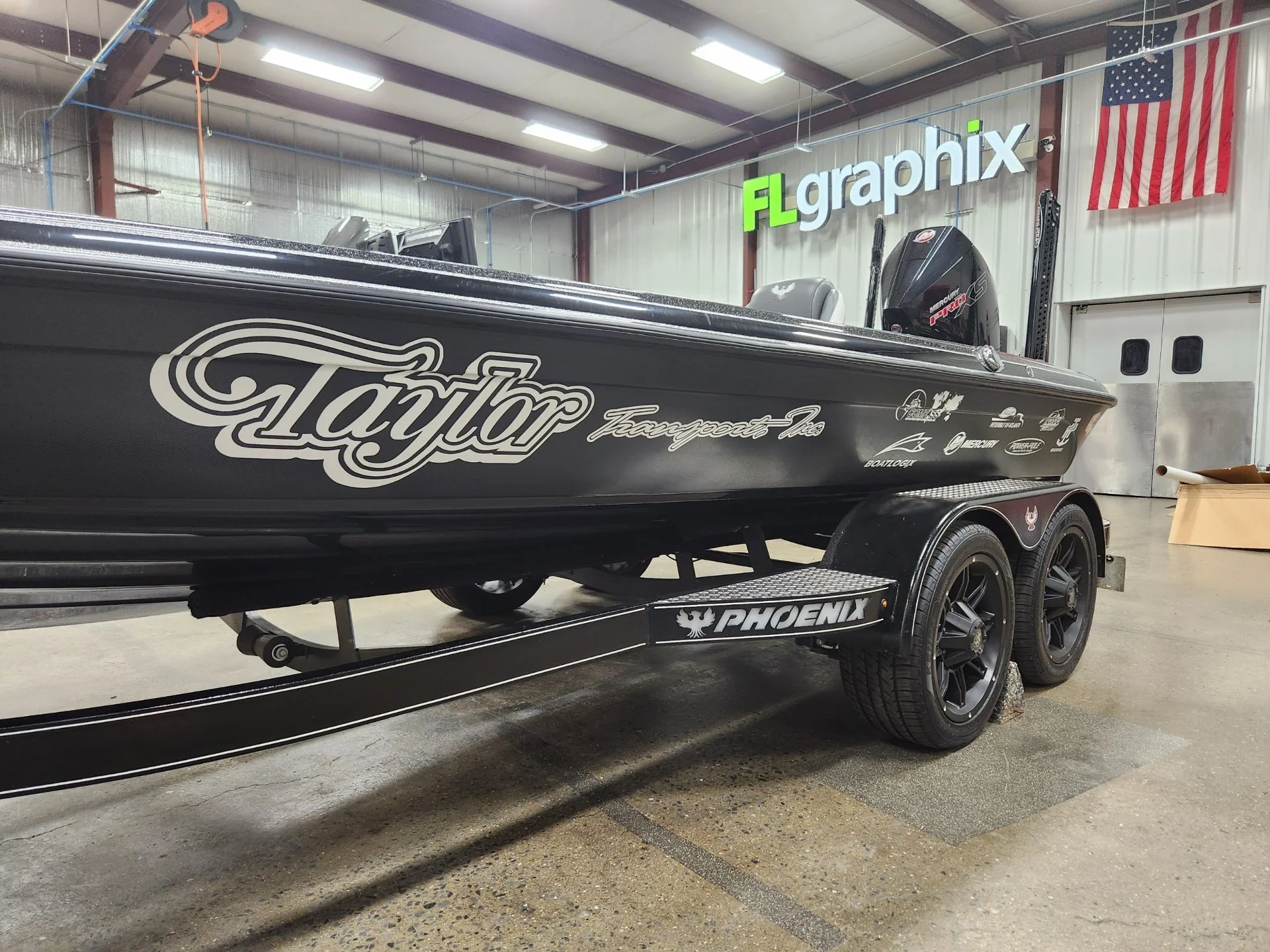 Black bass boat with Taylor and Phoenix logos, on a dual-axle trailer inside a warehouse with a high ceiling, American flag, and a sign reading 'FL graphix' on the wall.