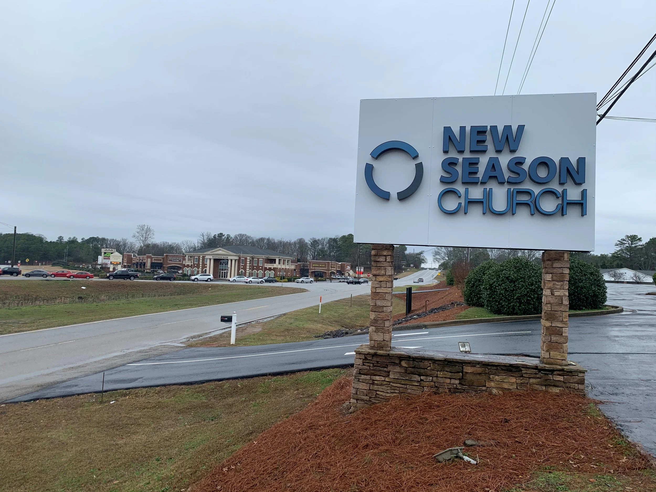 Signboard for New Season Church with brick pillars on a roadside, parking lot, shopping center in the background, overcast sky.