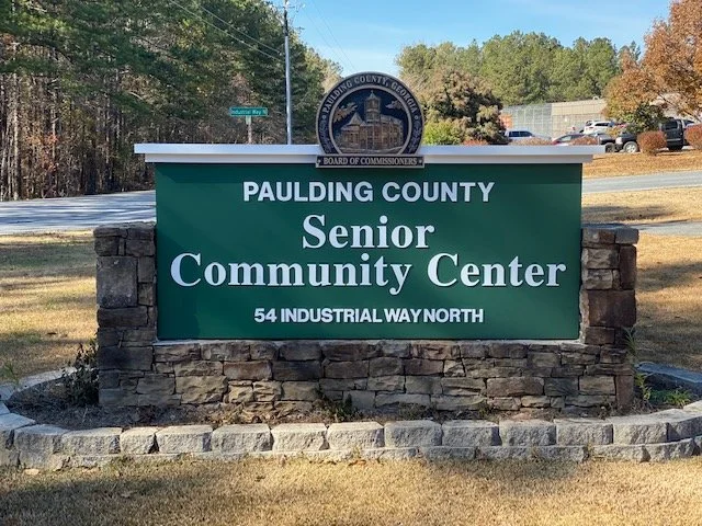 Sign for Paulding County Senior Community Center with address 54 Industrial Way North, located outdoors with trees and vehicles in the background.
