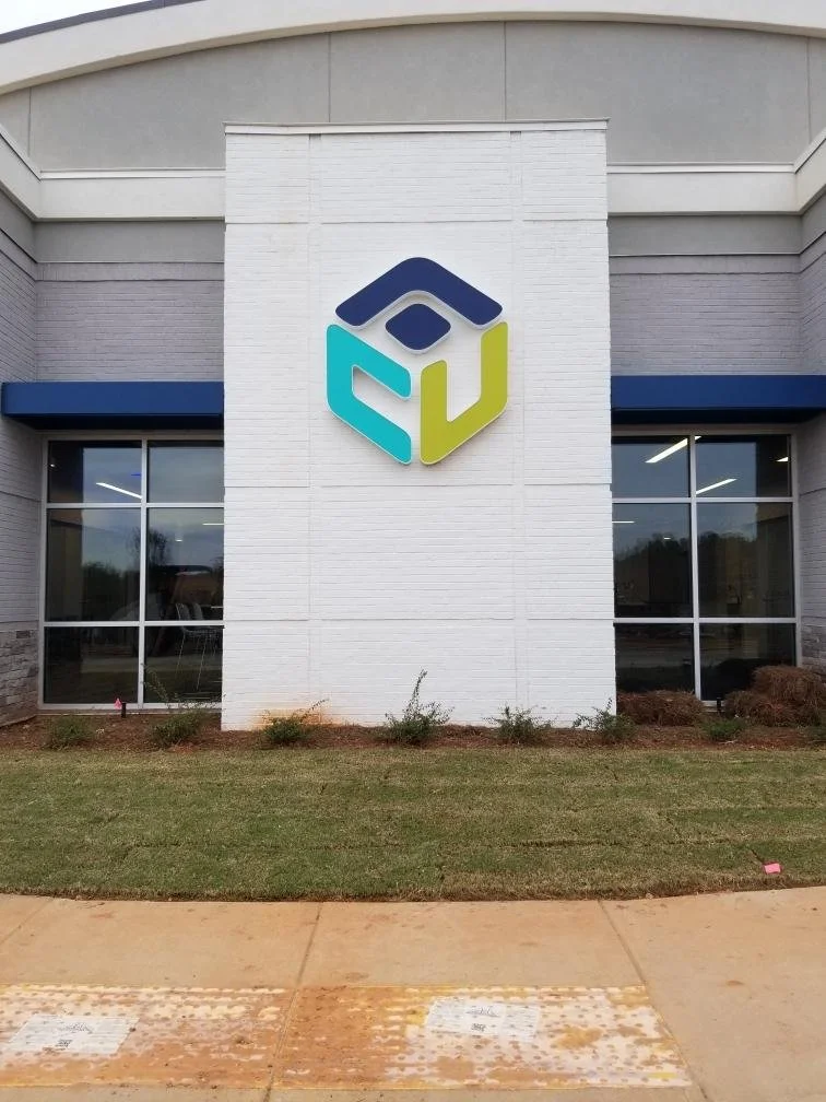 Building exterior with a colorful logo on a white brick wall, large glass windows on either side, and a small landscaped area in front.