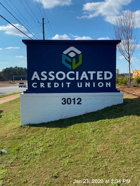Sign for Associated Credit Union with the address 3012 on a grassy area near a road, under a partly cloudy sky.